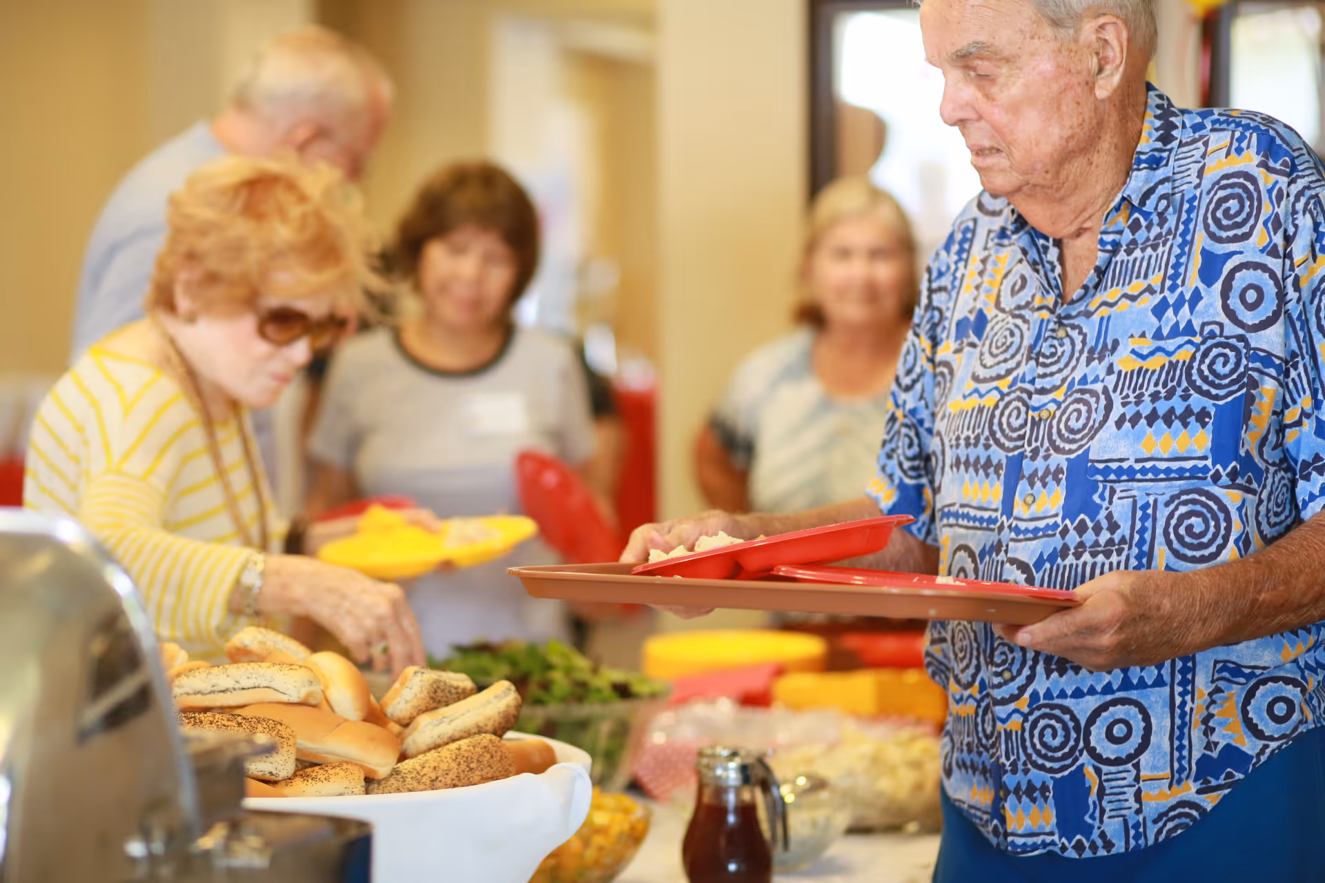 A group of elderly people serving themselves food from a buffet table with various breads, salads, and dishes in a communal dining setting.