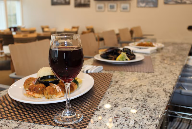 A glass of red wine and plated meals on a granite countertop in a dining area with chairs and tables in the background.
