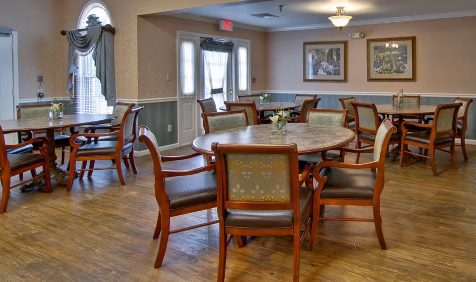 A dining room with several round and rectangular wooden tables surrounded by cushioned chairs. The room has wood flooring, beige walls with white trim, two framed pictures on the wall, and a door with windows letting in natural light. Small flower arrangements are placed on each table.