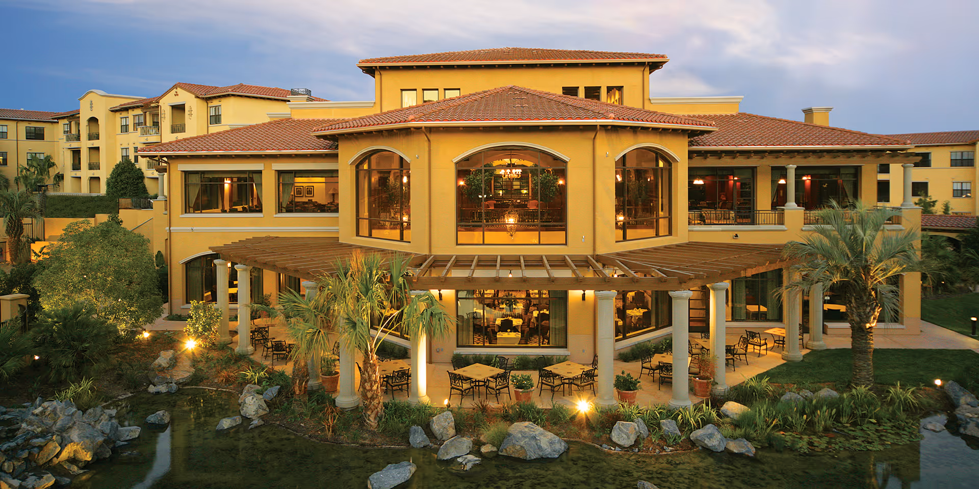 Exterior view of a large, multi-story senior living facility building with a yellow stucco facade and red tile roof. The building features large windows, a covered patio with tables and chairs, surrounded by landscaped greenery, palm trees, rocks, and a small pond in the foreground during dusk.