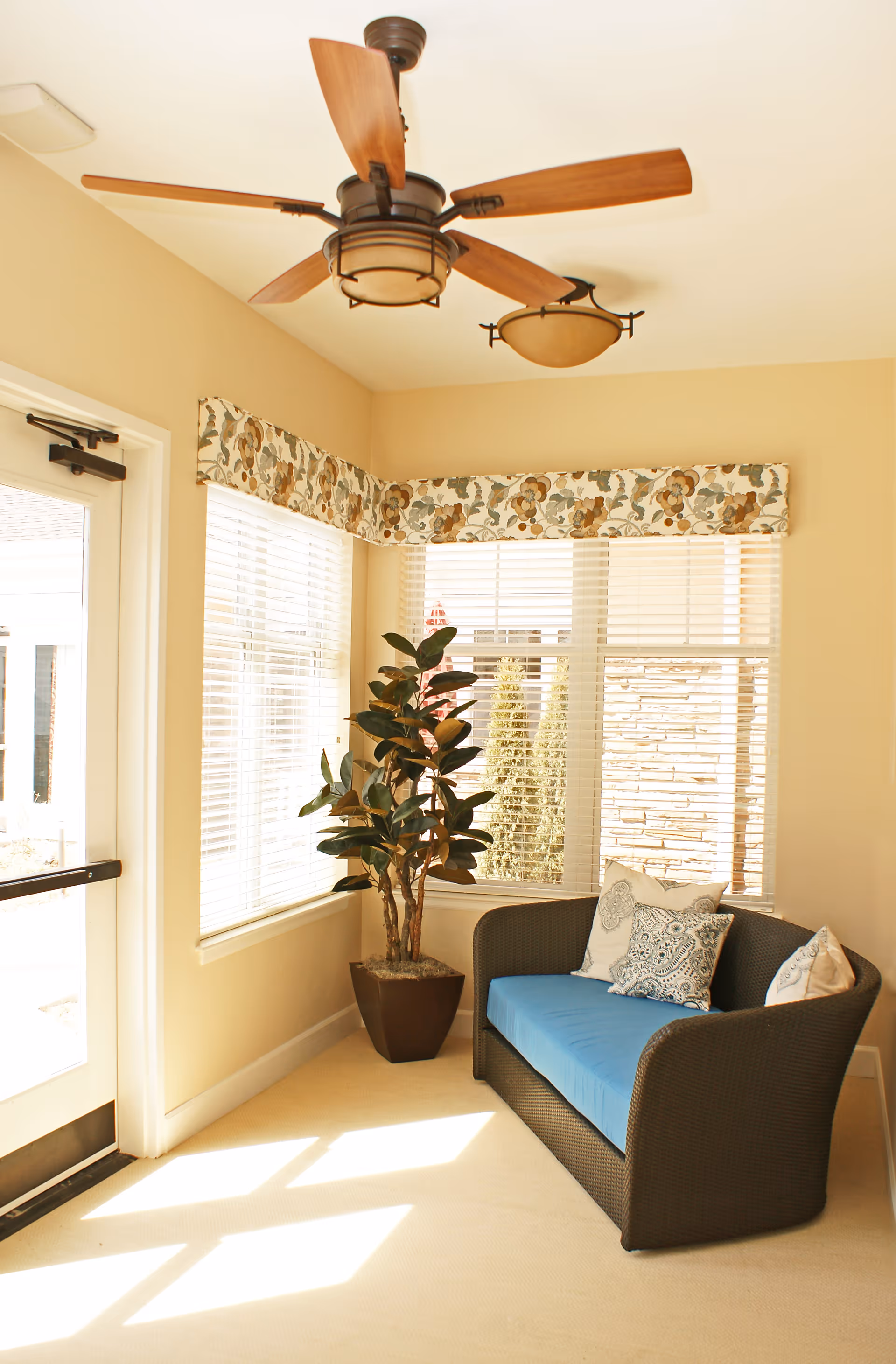 A bright corner of a room with large windows covered by white blinds and a floral valance. There is a dark wicker loveseat with a blue cushion and three decorative pillows. A potted plant is placed next to the loveseat. The ceiling has a wooden-blade ceiling fan and a light fixture. Sunlight streams through the windows onto the light-colored carpet.