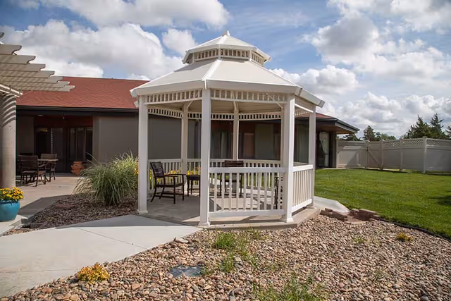 White gazebo with chairs in a landscaped courtyard beside a single-story building under a partly cloudy sky.