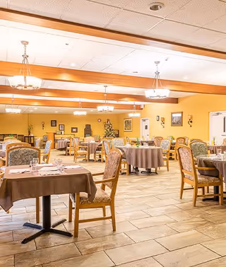 A spacious dining room with multiple round and square tables covered with brown tablecloths. Each table is set with glasses, plates, and napkins. The room has wooden beams on the ceiling, hanging light fixtures, and framed artwork on the yellow walls. There are upholstered chairs around the tables and a Christmas tree decorated in the background.