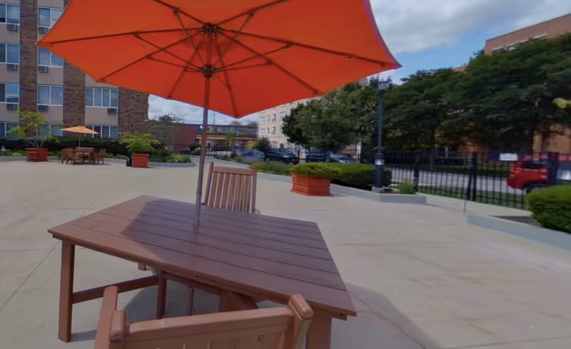 Outdoor patio area with wooden tables and chairs, each table shaded by a large orange umbrella. The patio is surrounded by buildings, greenery, and a black metal fence.