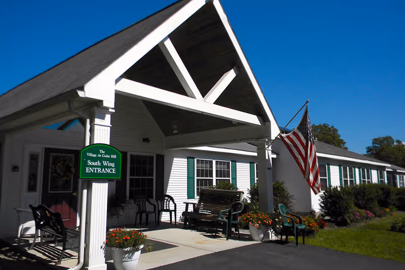 Exterior view of the Village At Cedar Hill Inc South Wing entrance with a covered porch, white building with green shutters, American flag, outdoor seating, and flower pots under a clear blue sky.