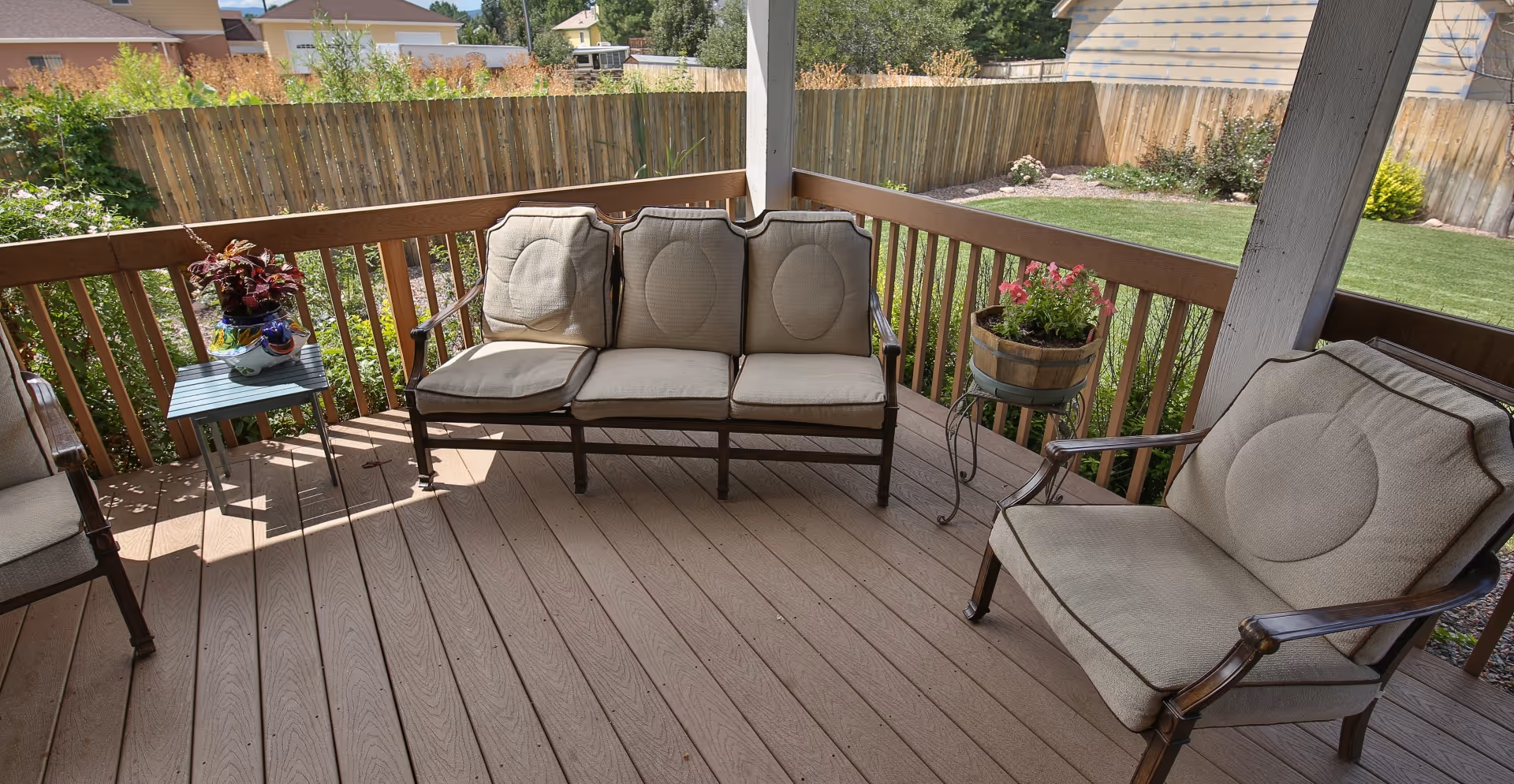 Covered outdoor patio area with cushioned seating including a three-seat sofa and a single armchair, a small side table with potted plants, wooden railing, and a fenced backyard with grass and shrubs.
