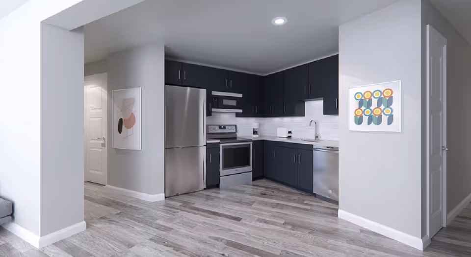 Modern kitchen with dark cabinets, stainless steel refrigerator, oven, microwave, and dishwasher. Light gray walls with two framed abstract artworks, and wood-look flooring.