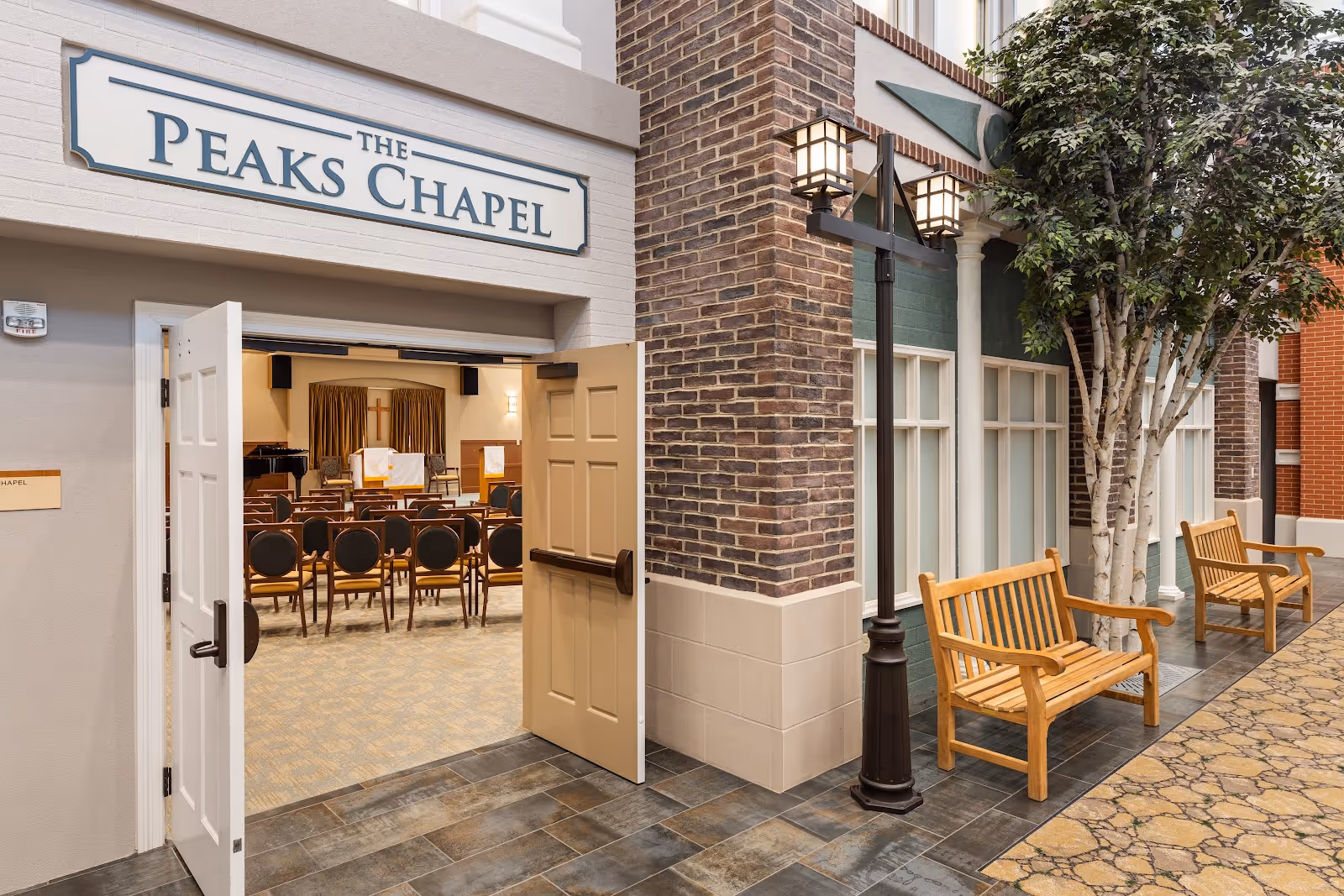 Entrance to The Peaks Chapel with open double doors revealing rows of chairs and a podium inside. Outside the chapel, there are two wooden benches, a streetlamp, and a tree along a tiled walkway with brick and painted walls.