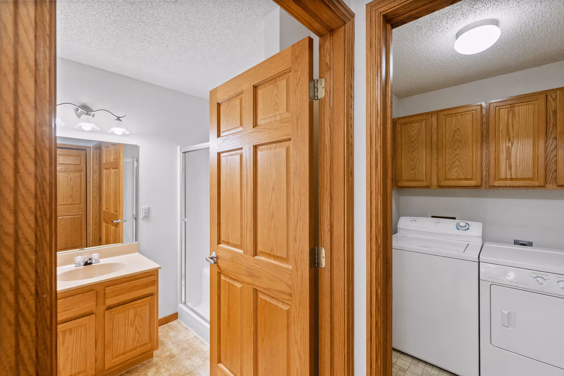 Interior view showing a bathroom with a wooden vanity, sink, mirror, and a shower stall on the left, and a laundry room with a washing machine, dryer, and wooden cabinets on the right, separated by wooden door frames.