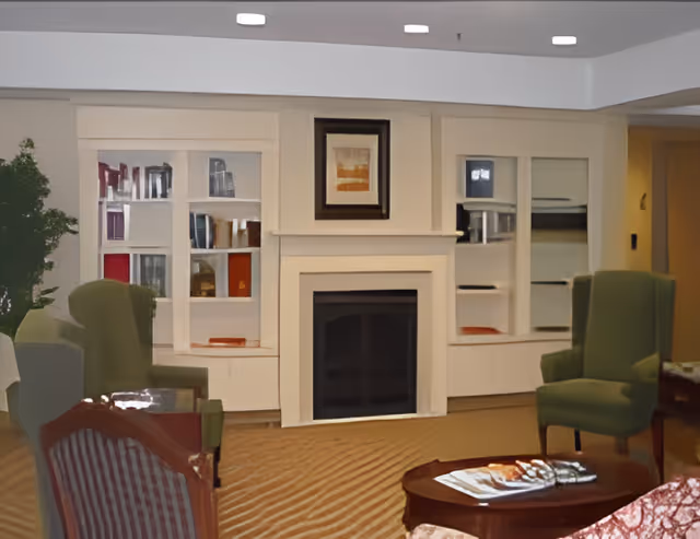Cozy assisted-living common room with a central fireplace, built-in shelving, armchairs, and a coffee table with magazines.