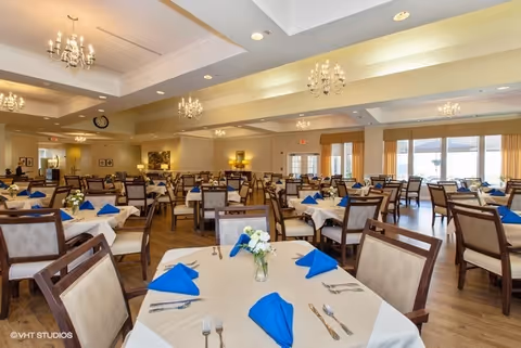 Spacious formal dining room with multiple tables set with white linens, blue napkins, floral centerpieces, and chandeliers.