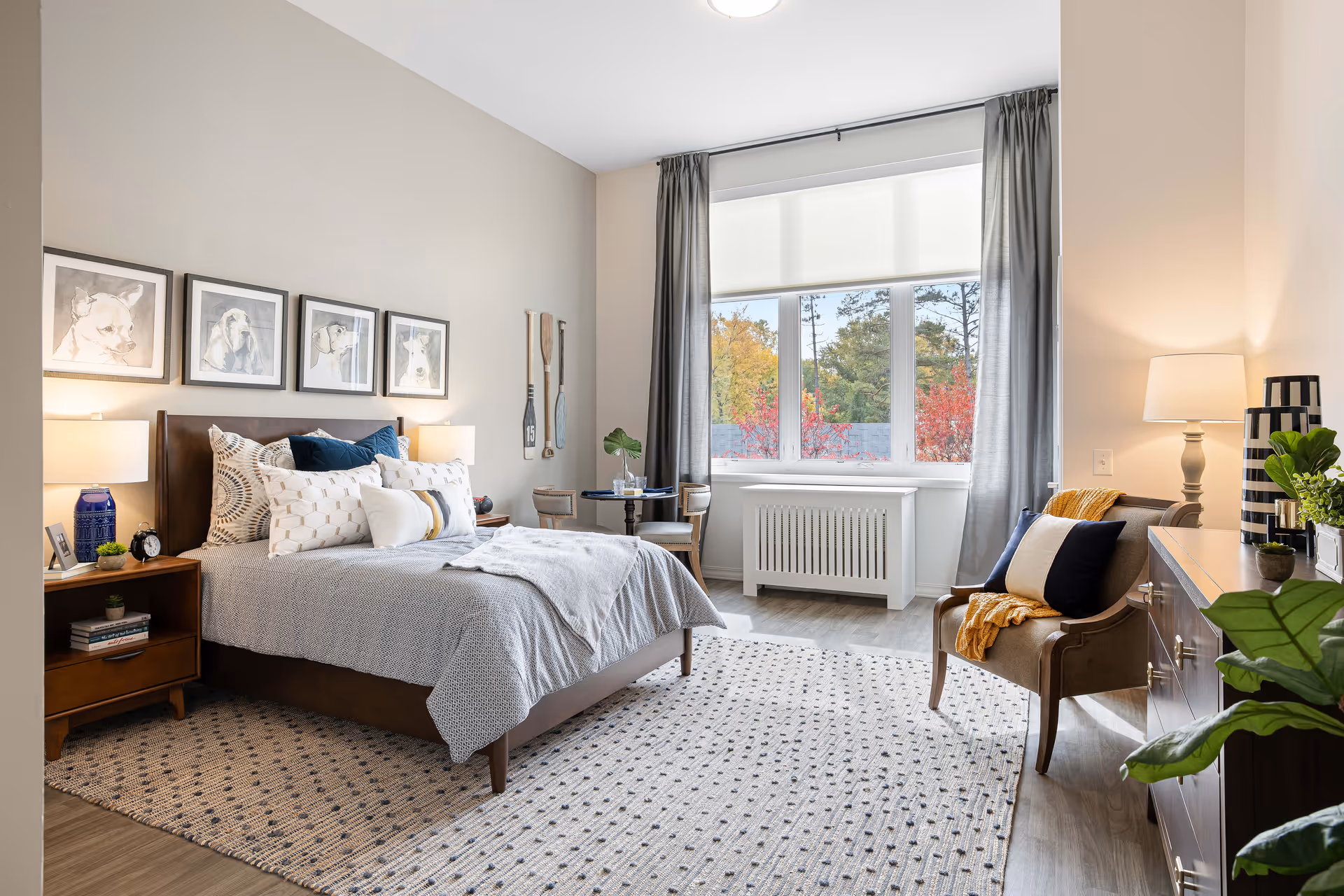 A bright and cozy bedroom featuring a large bed with multiple pillows and a gray bedspread. The room has a patterned rug, two bedside tables with lamps, framed dog artwork on the wall, and decorative paddles. A window with gray curtains lets in natural light, and there is a small round table with two chairs near the window. A comfortable armchair with a throw blanket and pillows sits next to a wooden dresser with plants and a lamp.
