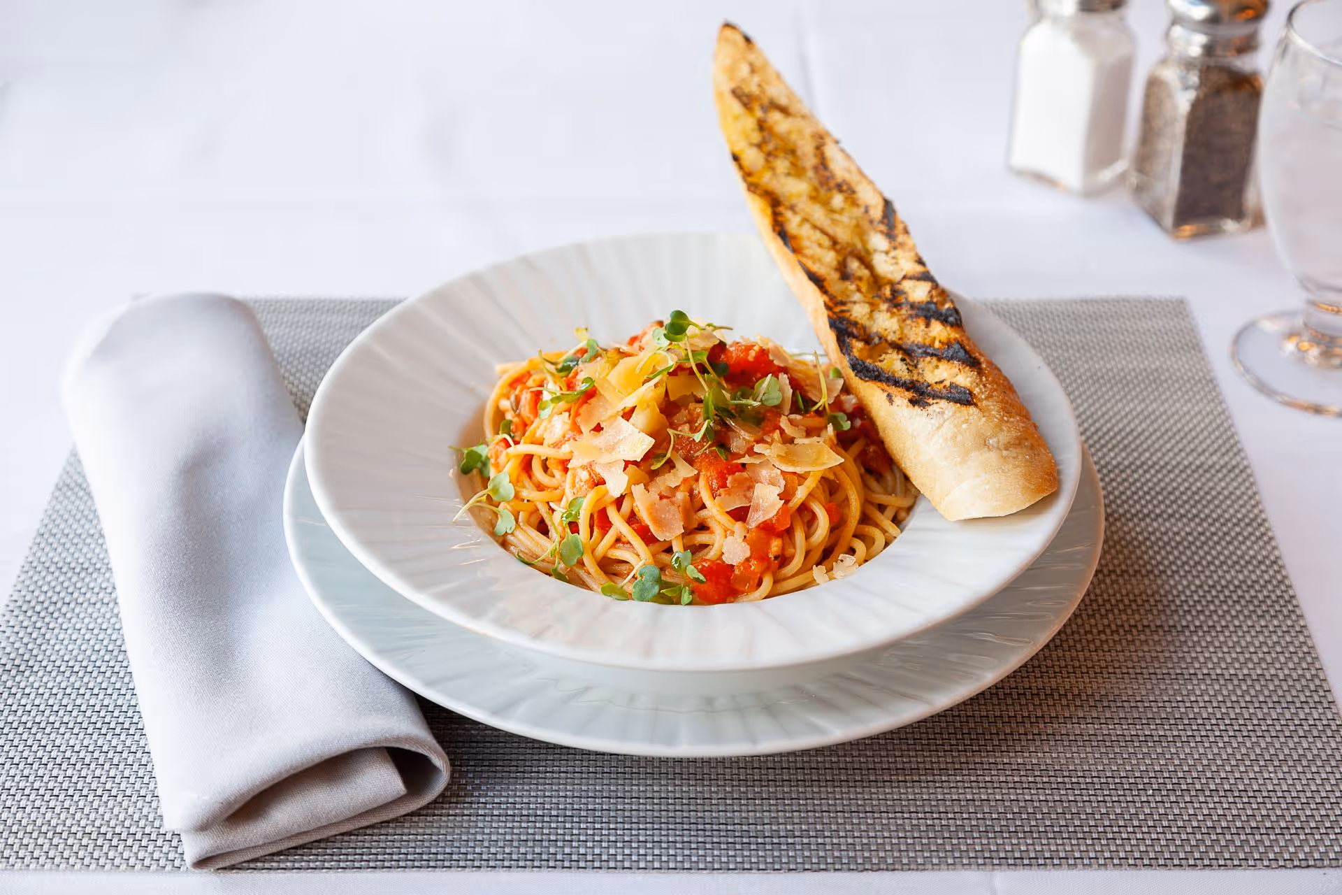 A bowl of spaghetti topped with tomato sauce, shaved cheese and microgreens, served with a grilled baguette on a white plate and gray placemat next to a folded napkin.