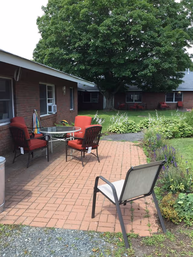 Outdoor courtyard patio with a glass table surrounded by red cushioned chairs on a brick paver area beside a low brick building and a large shade tree.