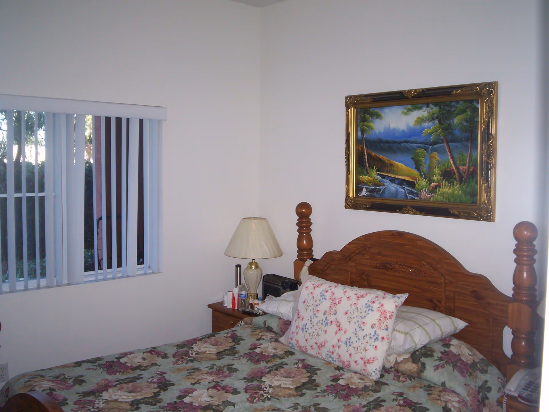 A bedroom with a wooden bed frame and floral bedspread. There are two pillows on the bed, one with a floral pattern and one with a checkered pattern. A wooden nightstand next to the bed holds a lamp with a cream-colored shade, a radio, and some small items. A window with vertical blinds is on the left wall, and a framed painting of a landscape with trees and a river hangs above the bed.