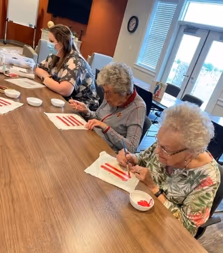 Three women sitting at a long wooden table engaged in a painting activity. Two elderly women are painting red stripes on white paper towels, while a younger woman wearing a face mask assists them. The room has large windows and a clock on the wall.