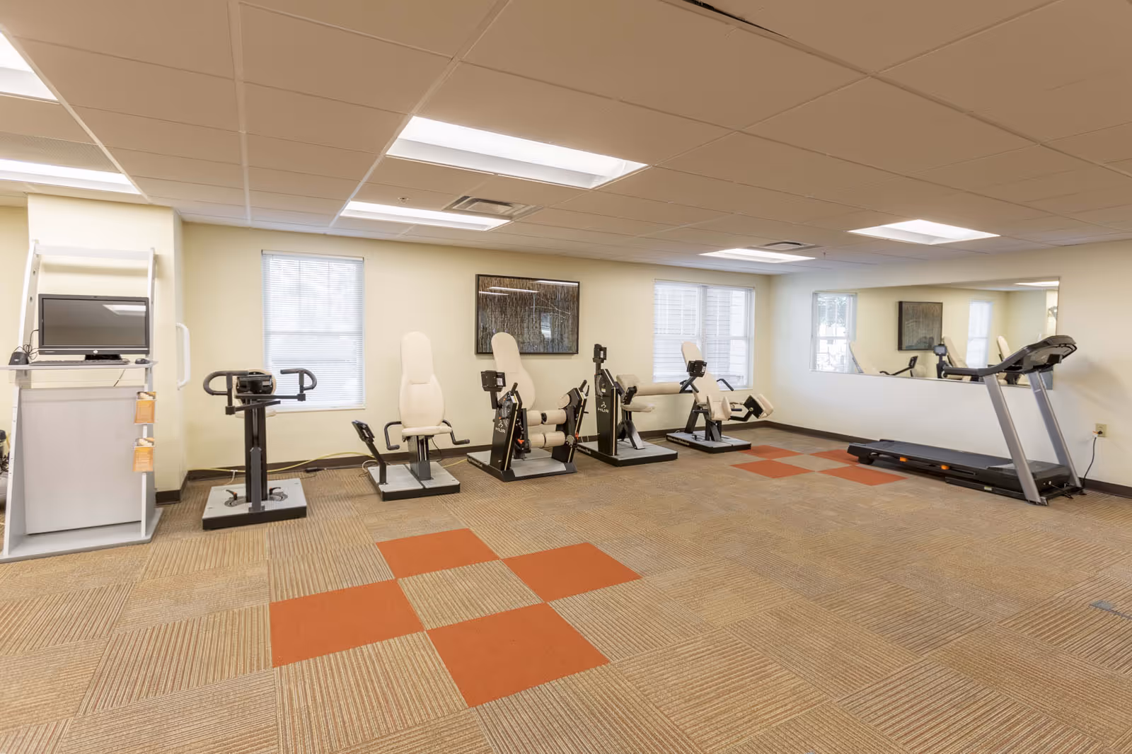 A well-lit senior exercise room with several fitness machines, a treadmill, windows, and a mirrored wall.