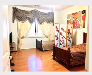 Spacious assisted-living bedroom with two wooden twin beds, a decorative folding screen, TV, and a large window with draped curtains.