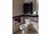 A small bathroom with a white toilet, a white sink, a wall-mounted black cabinet, and a towel rack holding two towels. The walls are tiled halfway up with white tiles and a dark border tile.