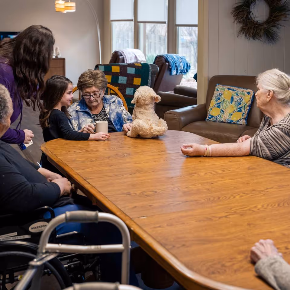 A group of elderly women and a young girl sitting around a wooden table in a cozy living room. One elderly woman is holding a mug, and a stuffed dog toy is placed on the table. The room has comfortable chairs with colorful blankets and pillows, and a large window in the background.