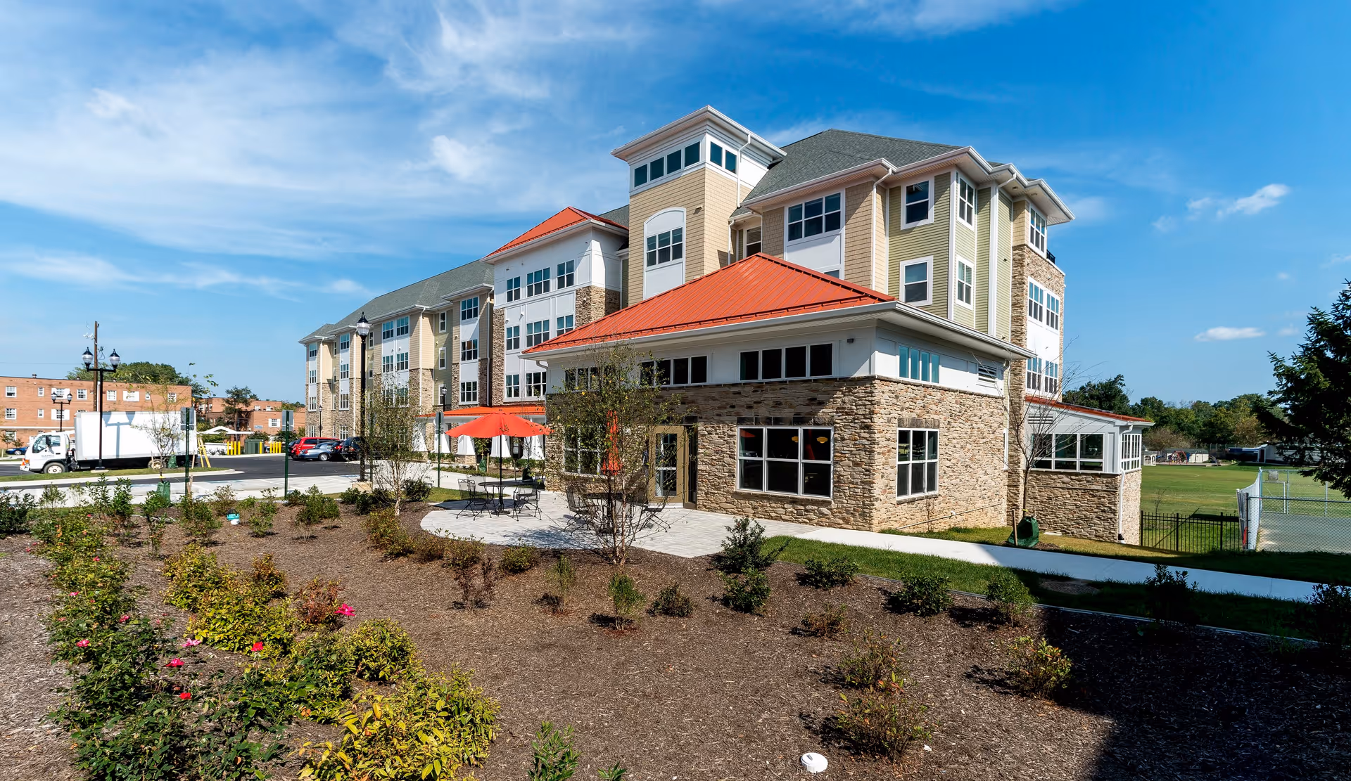 Exterior view of a multi-story senior living facility with beige and stone facade, red roofs, and large windows. There is a landscaped garden area with small bushes and flowers in the foreground, a patio with tables and red umbrellas, and a parking area with cars and a truck. The sky is clear with a few clouds.
