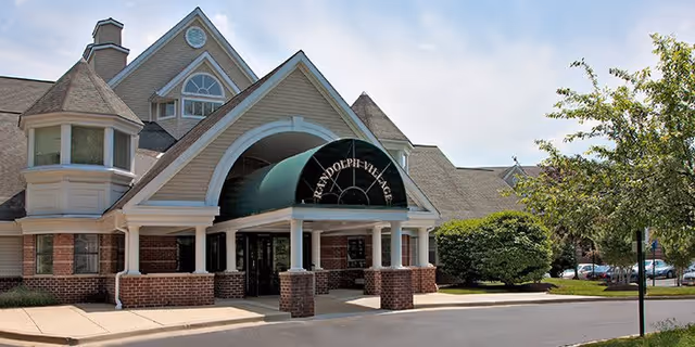 Front exterior view of Randolph Village facility showing a large entrance with a covered driveway, brick and beige siding, multiple peaked roofs, and surrounding greenery including trees and bushes.