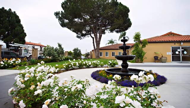 Outdoor garden area at Arroyo Vista Nursing Center featuring a black tiered fountain surrounded by purple and yellow flowers, white blooming bushes, a large tree, and a building with a red-tiled roof in the background.