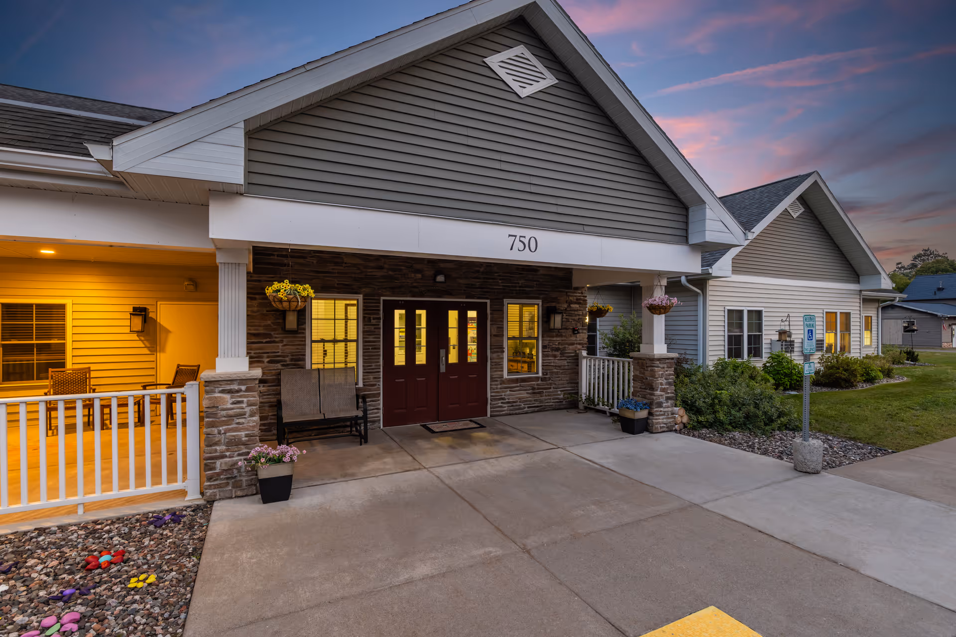 Front entrance of a senior living building with a covered porch, double doors, hanging planters, and the address number 750 at dusk.