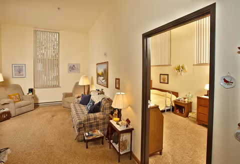 Cozy living room with a sofa and armchairs, lamps and side tables, and an open doorway revealing a bedroom.
