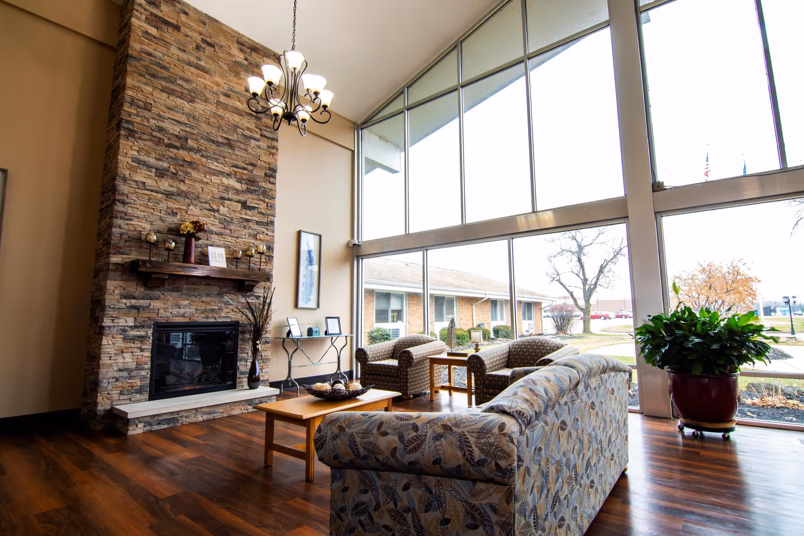 A bright and spacious living room area with large floor-to-ceiling windows letting in natural light. The room features a stone fireplace with a wooden mantel decorated with small items, a chandelier hanging from the ceiling, a patterned sofa, two armchairs, a wooden coffee table, and a large potted plant near the windows. Outside, a building and trees are visible.