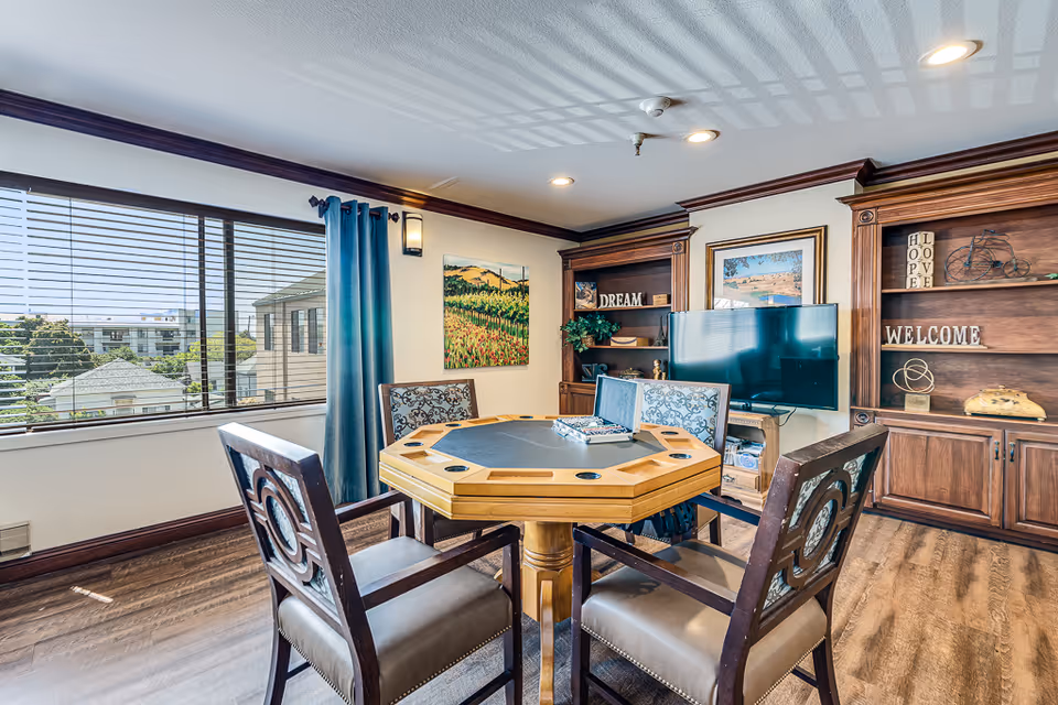 A bright and cozy common area in a senior living facility featuring a wooden octagonal table with four cushioned chairs around it. The room has large windows with blinds and blue curtains, wooden bookshelves with decorative items and signs that say 'DREAM' and 'WELCOME', a flat-screen TV, and framed artwork on the walls. The floor is wood, and the ceiling has recessed lighting.