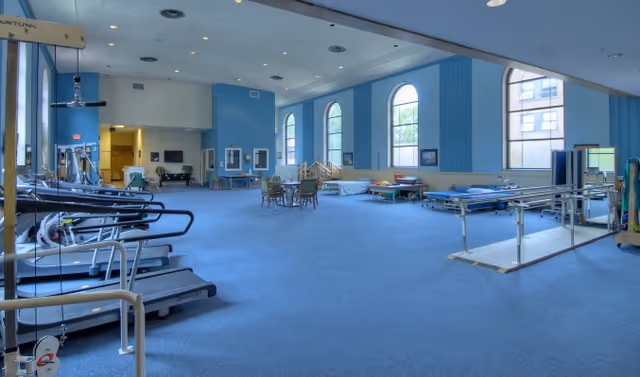 A spacious rehabilitation or physical therapy room with blue carpet and walls, large arched windows letting in natural light, several treadmills on the left side, parallel bars for walking exercises on the right, and various therapy equipment and tables scattered throughout the room.