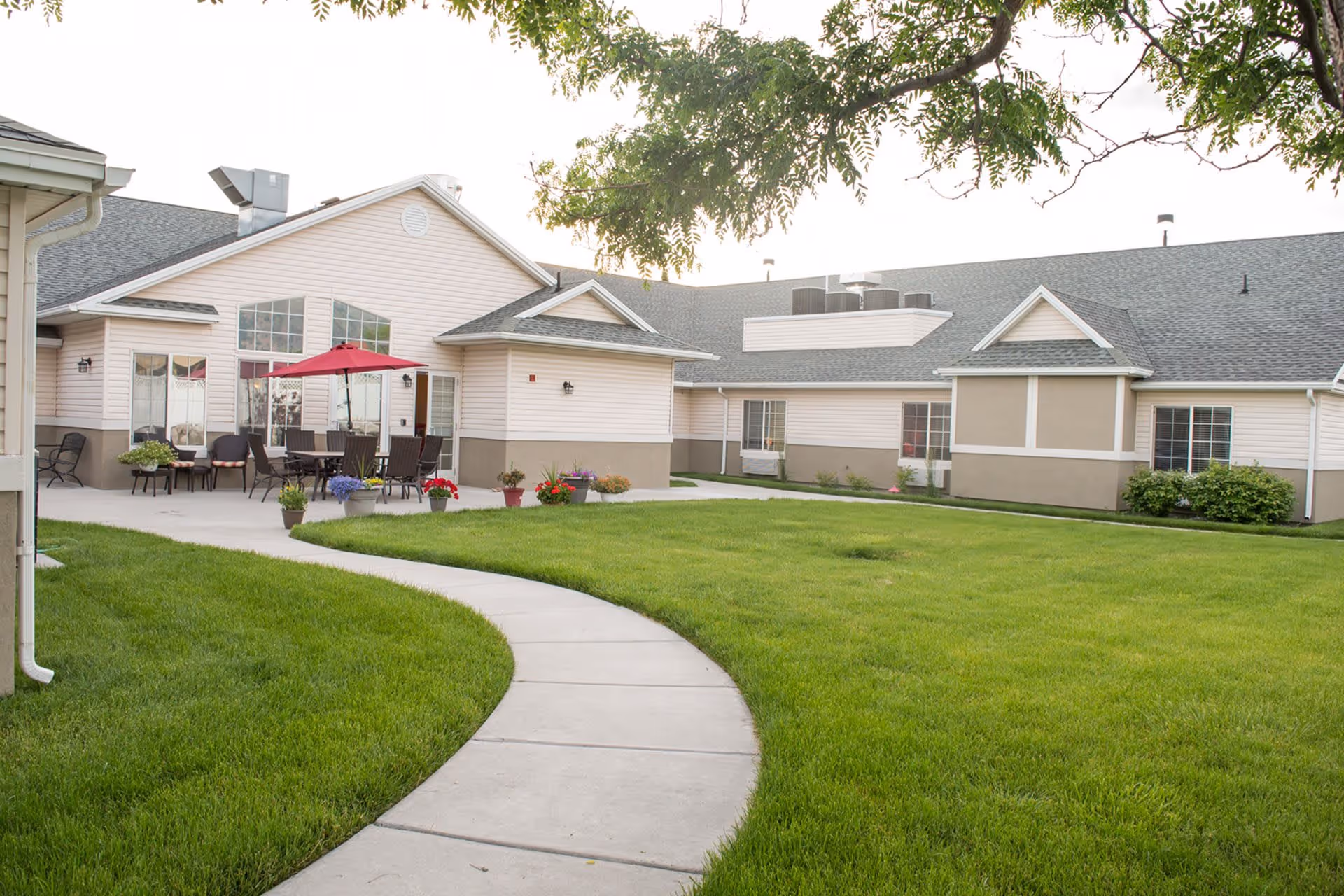 A well-kept courtyard with a curved concrete walkway, green lawn, patio seating and a red umbrella in front of a single-story assisted living building.