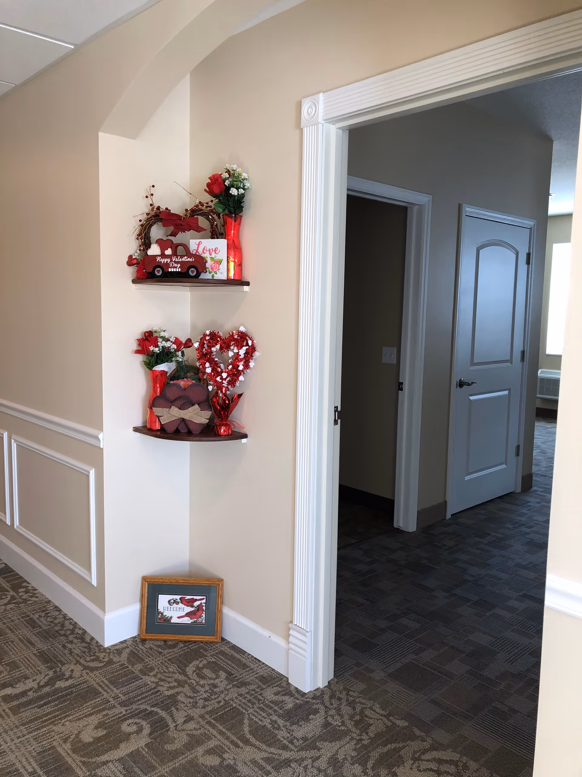 Interior corner of a senior living facility hallway decorated with Valentine's Day themed items including red vases with flowers, heart-shaped decorations, and a small framed picture on the floor. The hallway has beige walls, white trim, and patterned carpet. Two open doorways lead to other rooms.
