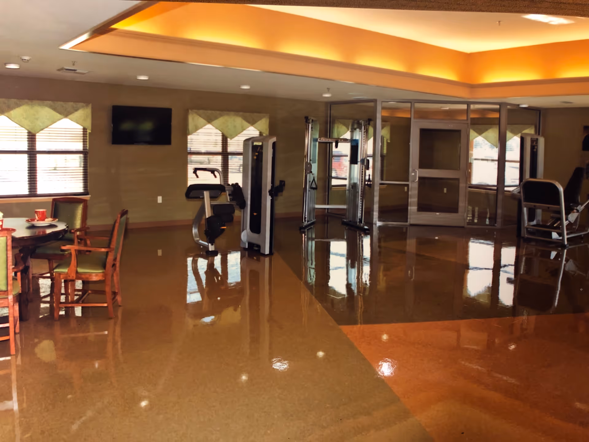 Interior view of a senior living facility room with exercise equipment including weight machines and a stationary bike. There is a round table with chairs on the left side, windows with green valances, a wall-mounted TV, and a glass door leading to another area. The floor is shiny and reflective with a brown and orange pattern, and the ceiling has recessed lighting with a warm glow.