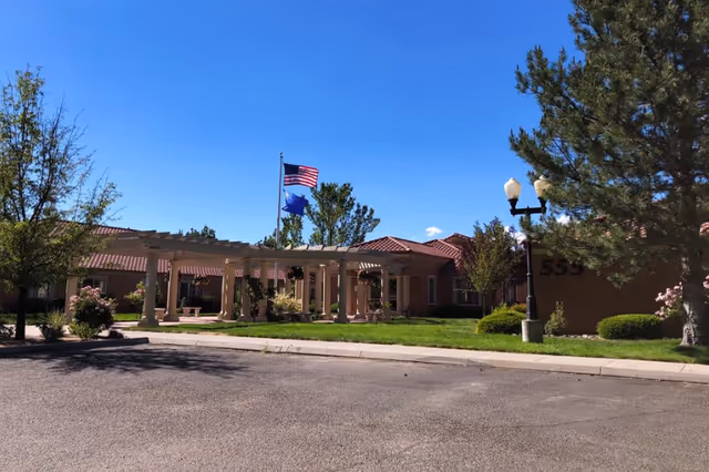 Exterior view of Alta Skilled Nursing And Rehabilitation Center showing a single-story building with a red-tiled roof, a covered entrance supported by columns, two flagpoles with the American and another flag, trees, bushes, and a clear blue sky.