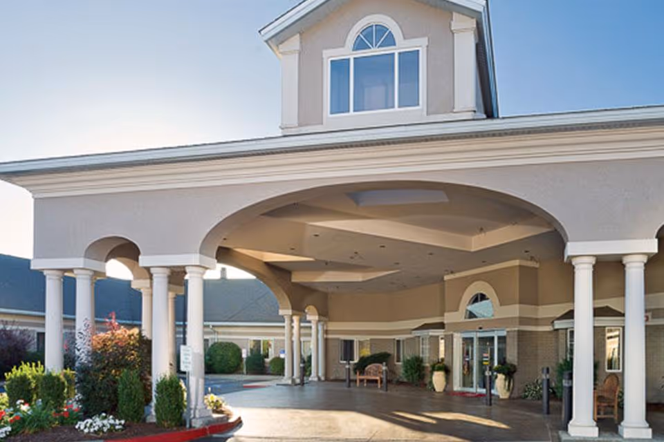 Entrance of a senior living facility with a covered drop-off area supported by white columns, landscaped greenery, and a clear sky in the background.