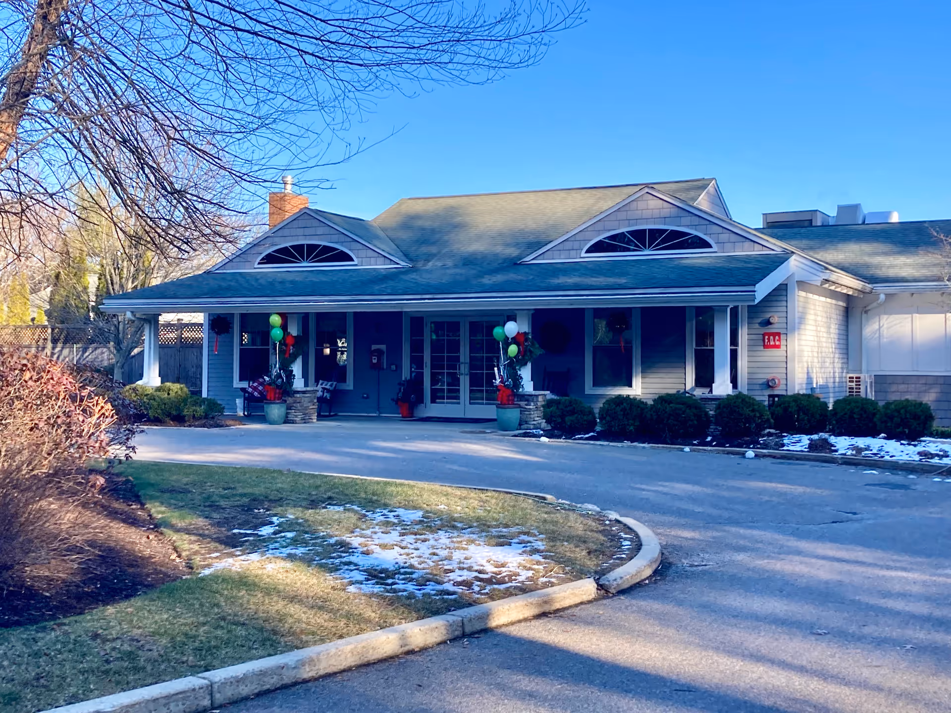 Exterior view of a single-story building with a covered entrance, decorated with balloons and holiday wreaths. The building has a green roof, beige siding, and a driveway leading up to the entrance. There are some bushes and patches of snow on the ground under a clear blue sky.