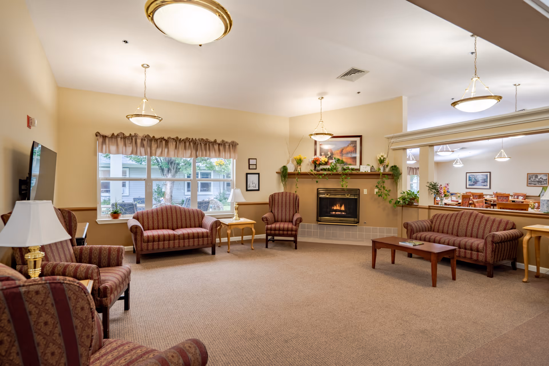 A cozy senior living facility common area with patterned upholstered armchairs and sofas arranged around a coffee table and side tables with lamps. A fireplace with a mantle decorated with plants and flowers is on one wall, and large windows with beige curtains let in natural light. A large mirror reflects a dining area with tables and chairs in the background.