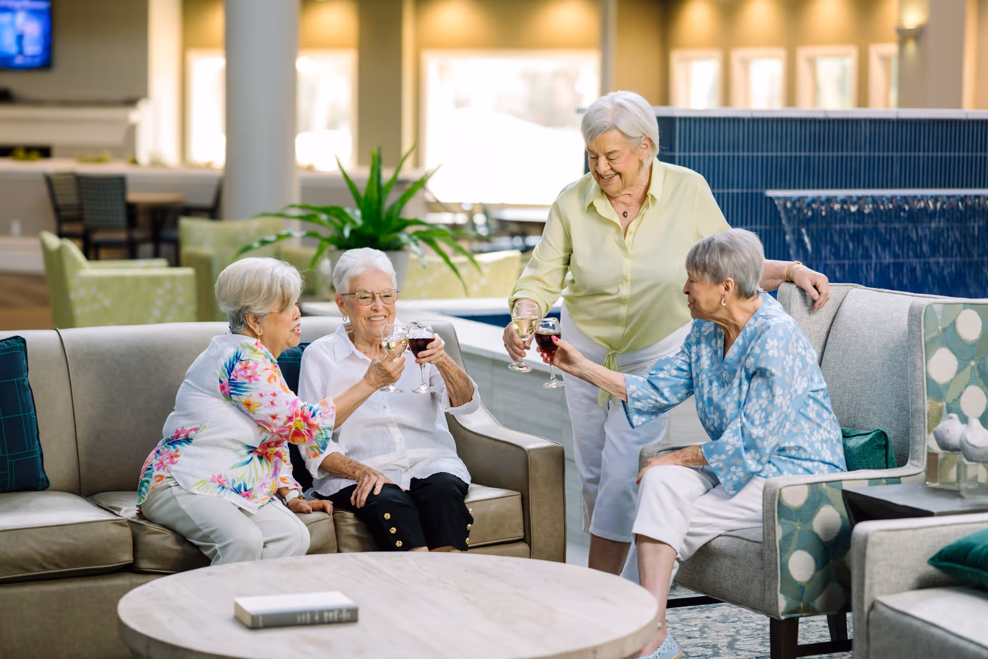 Four elderly women sitting and standing around a living room area, smiling and toasting with glasses of wine. The room has comfortable seating, a round wooden table, and a large plant in the background.