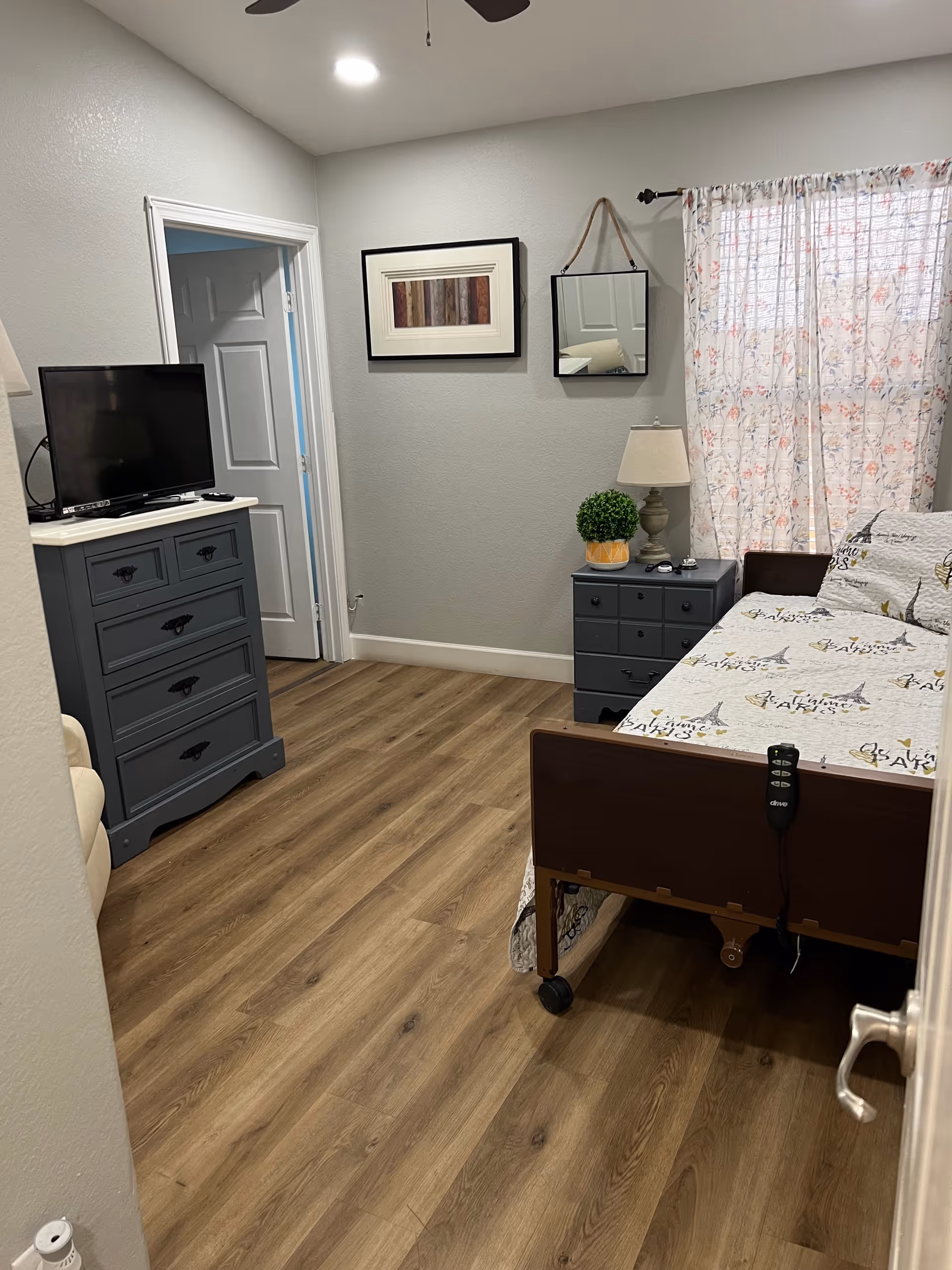 A senior living bedroom with wood flooring, a hospital-style bed with patterned bedding, a small bedside table with a lamp and a potted plant, a dresser with a flat-screen TV on top, a wall mirror, and a framed picture. A window with floral curtains lets in natural light.