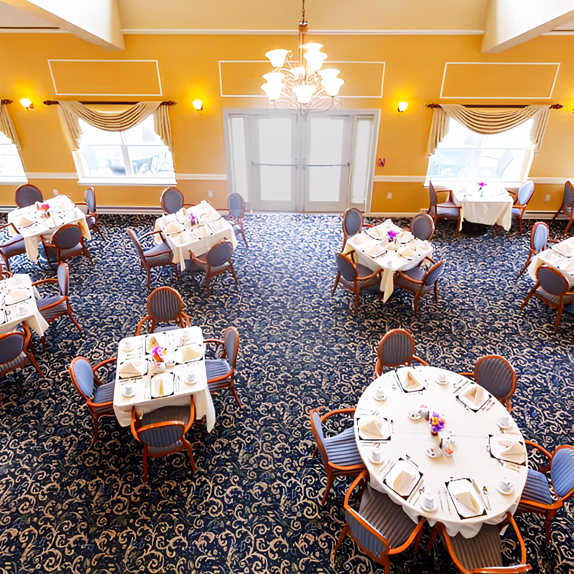 A dining room with multiple tables set with white tablecloths, napkins, plates, cups, and silverware. The room has patterned blue carpet, yellow walls, large windows with draped curtains, and a chandelier hanging from the ceiling.