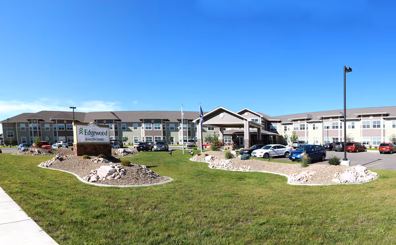 Wide exterior view of the Edgewood Assisted Living facility showing a large two-story building with multiple windows, a covered entrance, several parked cars, landscaped rock beds, green grass, and a clear blue sky.