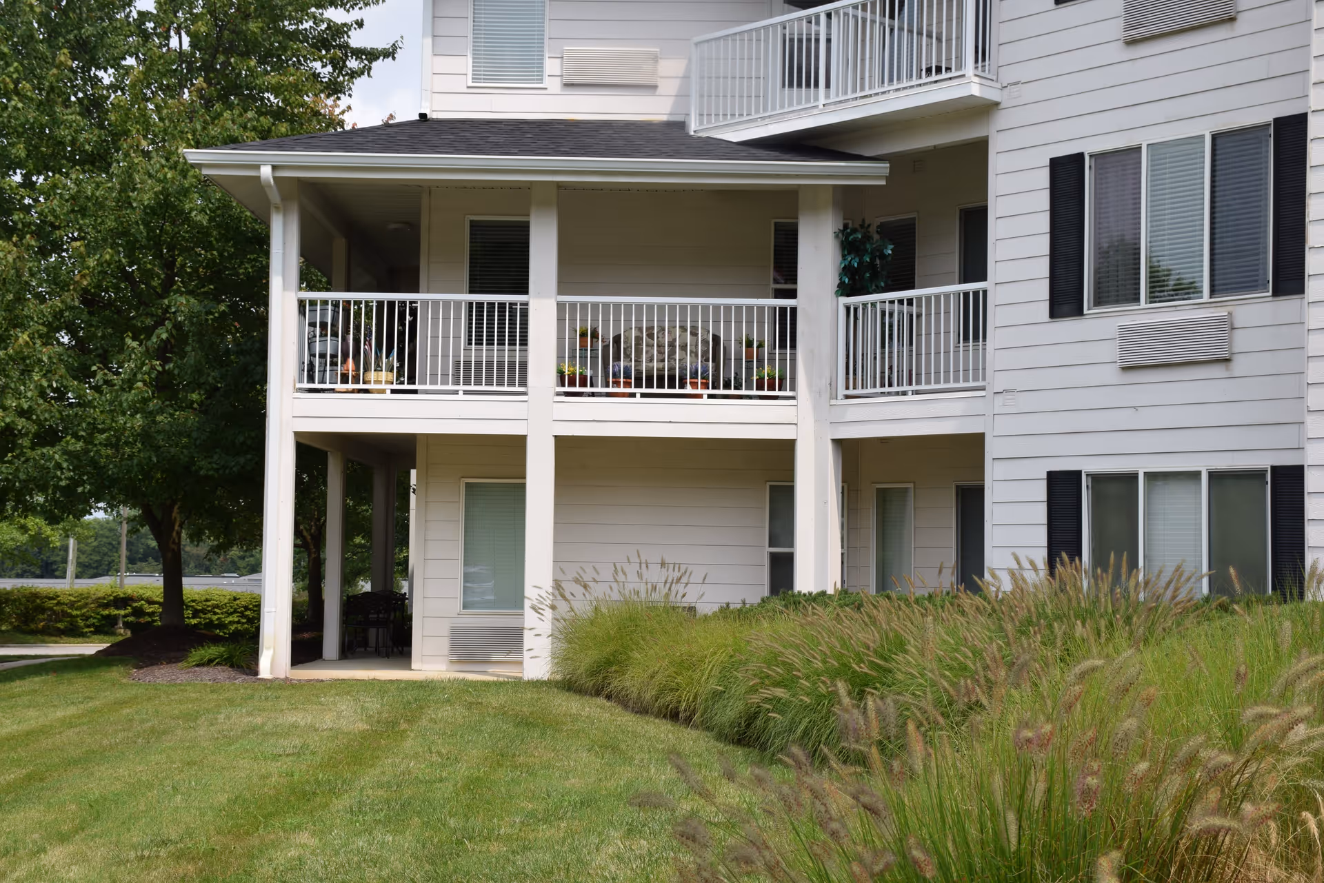 Exterior view of a two-story building with white siding and black window shutters. The building has a covered patio on the ground floor and a balcony on the second floor with potted plants. There is green grass and ornamental grasses in the foreground and trees in the background.