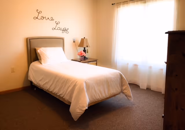 A simple and cozy bedroom with a single bed made with white bedding. There is a nightstand beside the bed with a lamp and a small vase of pink flowers. Above the bed, the words 'Love' and 'Laugh' are mounted on the wall in decorative script. A large window with sheer white curtains allows natural light to fill the room. The floor is carpeted and there is a wooden dresser partially visible on the right side.