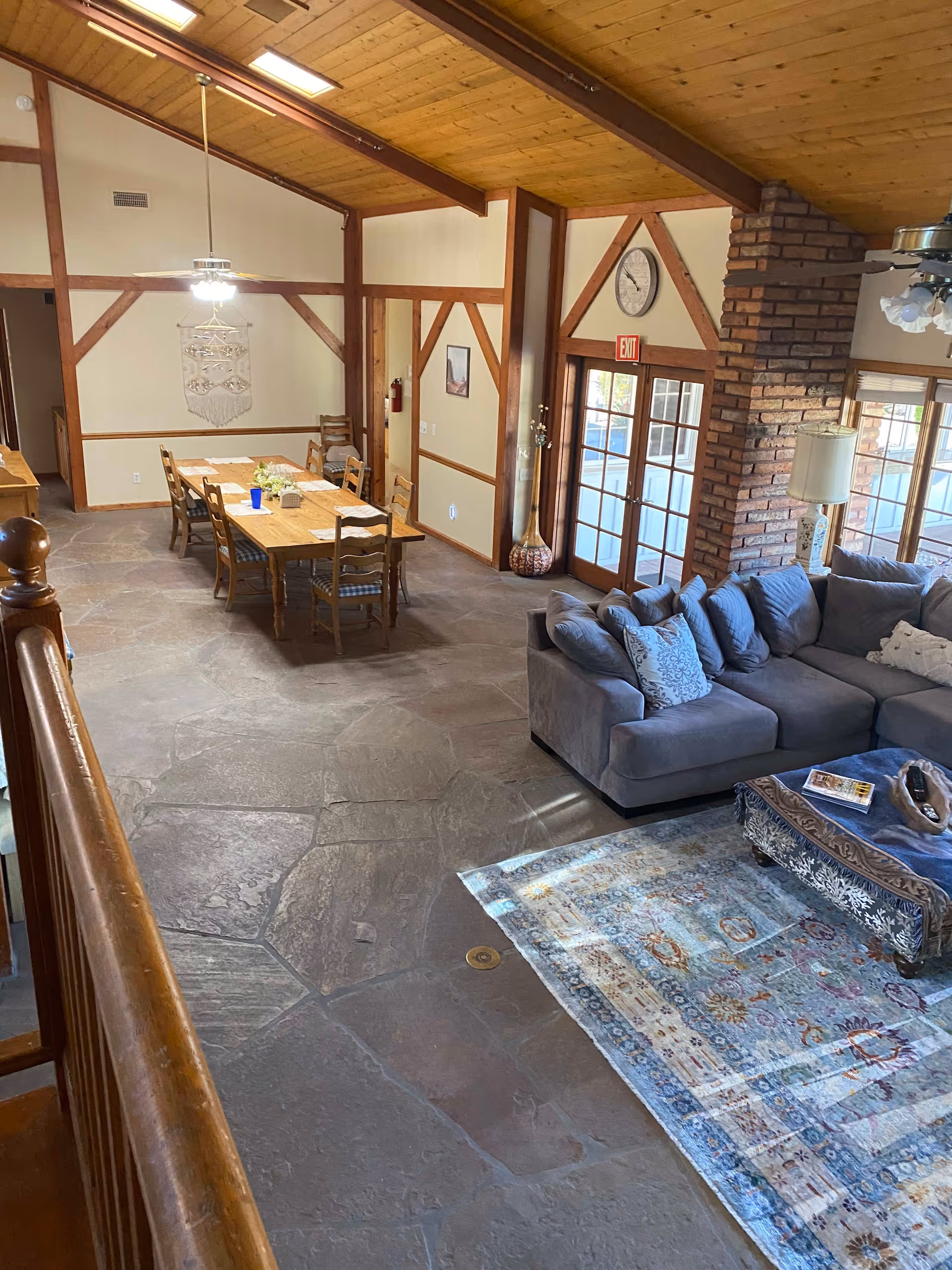 Interior view of a senior living facility common area with a wooden ceiling and beams. The room features a long wooden dining table with chairs, a gray sectional sofa with decorative pillows, a patterned area rug, and a coffee table. There are large windows and glass doors letting in natural light, a brick fireplace, ceiling fans, and a wall clock above the exit door.