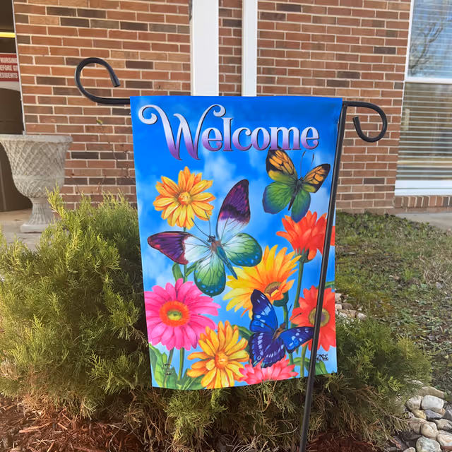 A colorful 'Welcome' garden flag featuring butterflies and flowers on a metal stand in front of a brick building and shrubs.