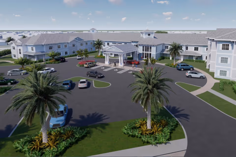 Exterior view of The Meridian at Punta Gorda Isles senior living facility showing a large building with multiple windows, a covered entrance with columns, several parked cars, palm trees, and landscaped greenery under a partly cloudy sky.