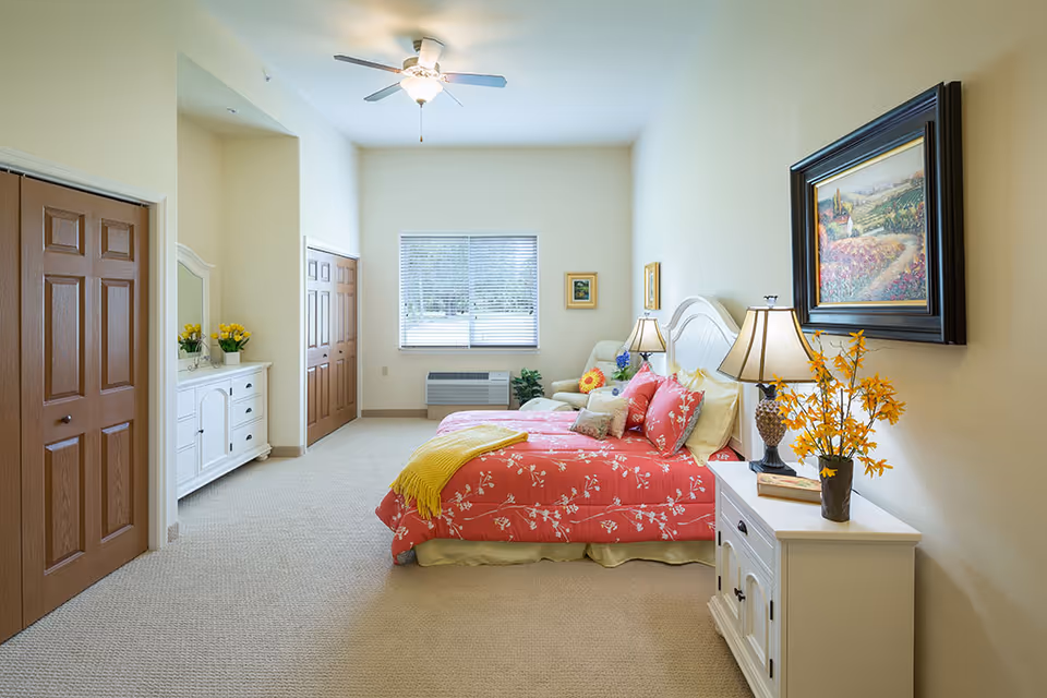 A bright and spacious bedroom with a large window, a ceiling fan, and beige walls. The room features a bed with a red floral bedspread and multiple pillows, a white nightstand with a lamp and a vase of yellow flowers, a white dresser with a mirror and yellow flowers, and two wooden closets with brown doors. A framed painting hangs on the wall above the nightstand.