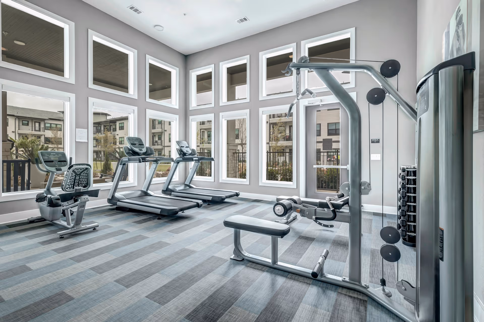 Bright fitness room with treadmills, a recumbent bike, and weight machine in front of a wall of large windows overlooking the complex.
